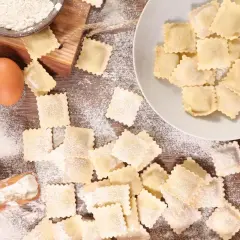 Ravioli fatti in casa sparsi su un tavolo di legno, con farina, uova e una ciotola di farina.