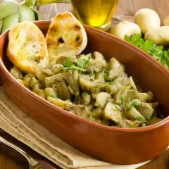 Un saporito piatto di carciofi in umido con patate, guarnito con pane tostato e prezzemolo fresco. Ingredienti freschi come carciofi, aglio e patate completano la scena.