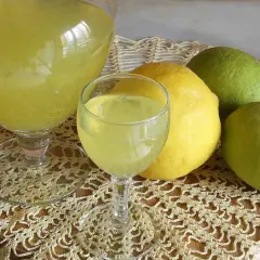 Limoncello ricetta originale Amalfi: bicchieri di liquore giallo brillante e limoni freschi, alcuni verdi, altri gialli, su un centrino di pizzo.