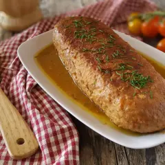 Un succulento polpettone in padella, dorato e profumato, adagiato su un letto di salsa e guarnito con erba cipollina. Accanto, una spatola in legno e pomodorini freschi.