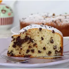 Una fetta di torta soffice con tante gocce di cioccolato, spolverata di zucchero a velo, su un piatto lilla con una forchetta.