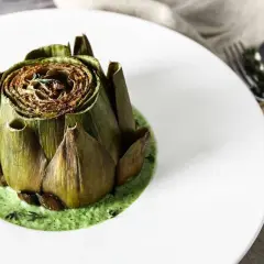 Carciofo intero cotto, adagiato su una vellutata crema di carciofi, guarnito con erbe aromatiche.