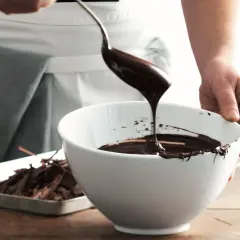 Mani che versano cioccolato fuso da un cucchiaio in una ciotola bianca, mostrando come sciogliere il cioccolato per dolci deliziosi.