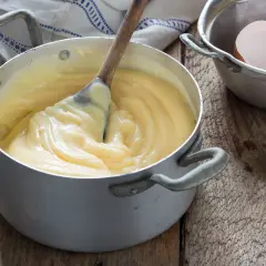 Crema pasticcera veloce in pentola, con cucchiaio di legno che mescola. Guscio d'uovo accanto, pronto per un dolce.
