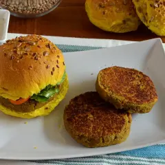 Hamburger di lenticchie dorato con semi, servito su un piatto bianco con due polpette aggiuntive e una ciotola di salsa.