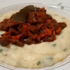 Piatto di purè cremoso con pezzetti di coratella di agnello stufata e una foglia di alloro.