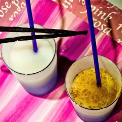 Due bicchieri con latte di riso, uno bianco e uno con spezie dorate, adornati da baccelli di vaniglia e cannucce blu su sfondo rosa.