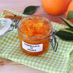 Barattolo di marmellata di arance fatto in casa, con arance fresche, foglie verdi e bastoncini di cannella sullo sfondo.