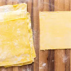 Fogli di pasta gialla, pronti per diventare deliziose lasagne fatte in casa. Un foglio singolo è posato a destra, mentre una pila di altri è a sinistra.