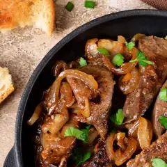 Fegato alla veneziana succulento in padella con cipolle caramellate e prezzemolo fresco, accompagnato da fette di pane tostato.