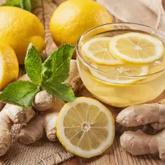 Una tazza di tisana zenzero e limone, con limoni freschi, zenzero e foglie di menta su un tavolo di legno.