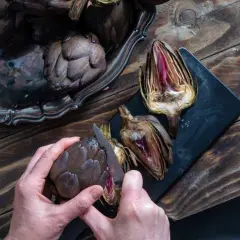 Mani che preparano carciofi su un tagliere scuro. Un carciofo intero viene tagliato con un coltello, mostrando il suo interno viola.