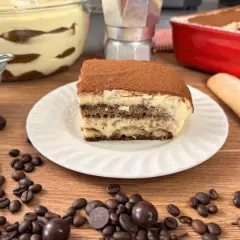 Una fetta di tiramisù con uova pastorizzate, cosparso di cacao, su un piatto bianco, circondato da chicchi di caffè e cioccolatini.