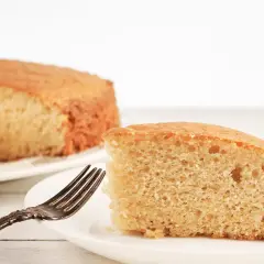 Una fetta di torta senza lievito, soffice e dorata, attende su un piatto bianco con una forchetta antica.