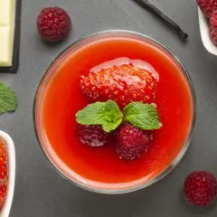 Un bicchiere di coulis di fragole, guarnito con fragola, lamponi e menta. Accanto, fragole fresche, cioccolato bianco, bacca di vaniglia e latte.