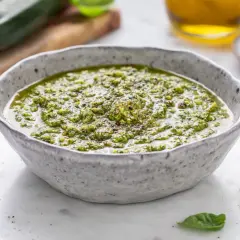 Pesto di zucchine cremoso in una ciotola, con zucchine fresche e pistacchi sullo sfondo.