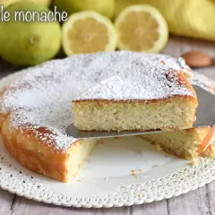 Torta delle monache soffice e profumata, cosparsa di zucchero a velo, con limoni e mandorle sullo sfondo.