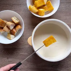 Una fetta di pane croccante viene intinta in una cremosa fonduta di parmigiano, accanto a ciotole di crostini.