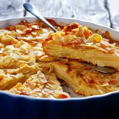 A slice of golden-brown apple cake with thin apple layers, topped with toasted almond slivers, is lifted from a blue baking dish.