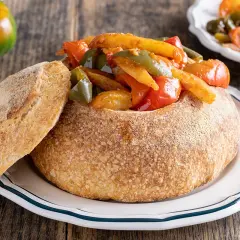 Un pane scavato ripieno di un saporito stufato di pipi e patate, con una fetta di pane croccante a lato.