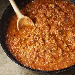 Un saporito ragù di carne in una padella, pronto per essere servito. Un cucchiaio di legno mescola la salsa rossa e densa.