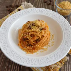 Un piatto di spaghetti con acciughe, pomodoro e pangrattato tostato, servito su un piatto bianco decorato, con un tovagliolo giallo e un bicchiere di pangrattato a lato.