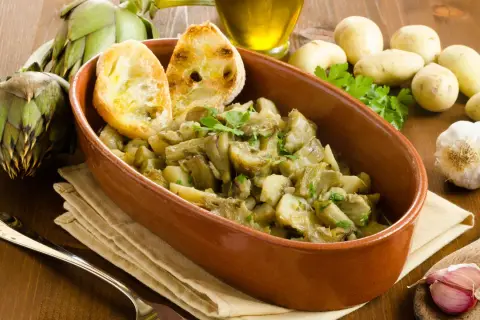 Un saporito piatto di carciofi in umido con patate, guarnito con pane tostato e prezzemolo fresco. Ingredienti freschi come carciofi, aglio e patate completano la scena.