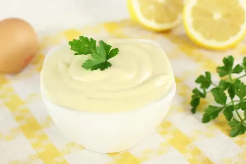 Una ciotola di cremosa maionese fatta in casa, guarnita con prezzemolo. Ingredienti freschi come uovo e limone sullo sfondo.
