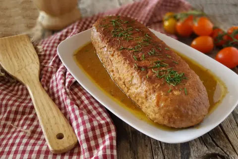 Un succulento polpettone in padella, dorato e profumato, adagiato su un letto di salsa e guarnito con erba cipollina. Accanto, una spatola in legno e pomodorini freschi.