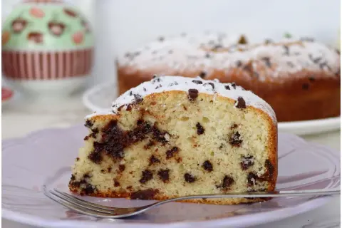 Una fetta di torta soffice con tante gocce di cioccolato, spolverata di zucchero a velo, su un piatto lilla con una forchetta.