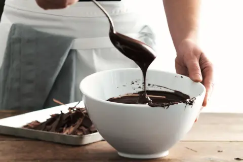 Mani che versano cioccolato fuso da un cucchiaio in una ciotola bianca, mostrando come sciogliere il cioccolato per dolci deliziosi.