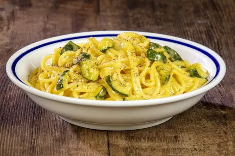 Spaghetti with zucchini and creamy sauce in a white bowl with blue trim on a wooden table.