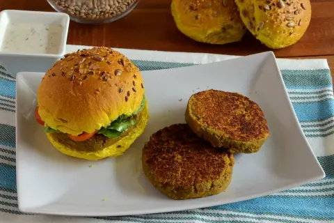 Hamburger di lenticchie dorato con semi, servito su un piatto bianco con due polpette aggiuntive e una ciotola di salsa.