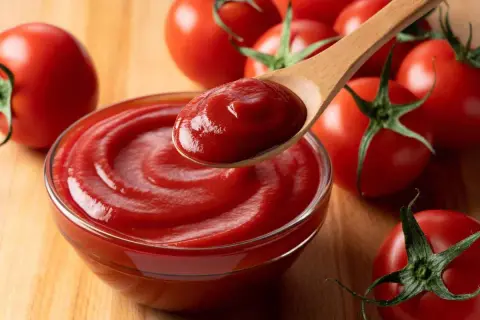 Un cucchiaio di cremoso ketchup fatto in casa, pronto per essere gustato, circondato da pomodori freschi su un tagliere di legno.