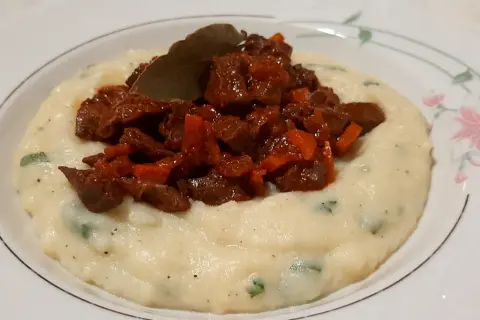 Piatto di purè cremoso con pezzetti di coratella di agnello stufata e una foglia di alloro.
