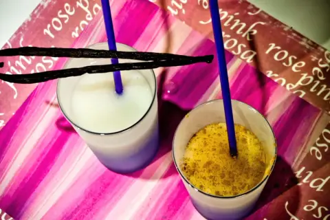 Due bicchieri con latte di riso, uno bianco e uno con spezie dorate, adornati da baccelli di vaniglia e cannucce blu su sfondo rosa.
