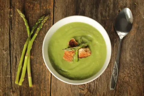 Crema di asparagi vellutata, guarnita con crostini e asparagi freschi, su un tavolo di legno rustico.