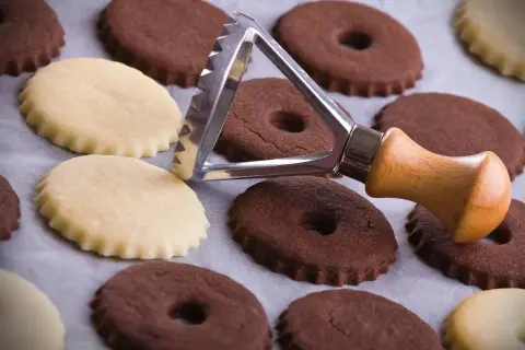 Biscotti al cacao e chiari, alcuni con foro centrale, pronti per essere infornati. Un tagliabiscotti con manico in legno è appoggiato su di essi.