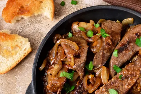 Fegato alla veneziana succulento in padella con cipolle caramellate e prezzemolo fresco, accompagnato da fette di pane tostato.