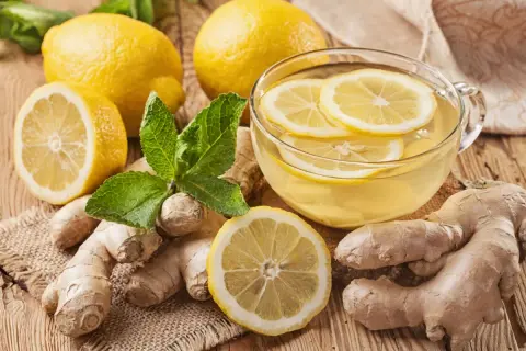 Una tazza di tisana zenzero e limone, con limoni freschi, zenzero e foglie di menta su un tavolo di legno.