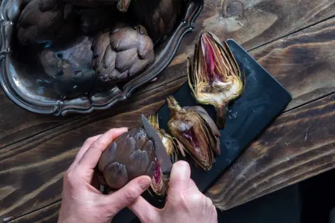 Mani che preparano carciofi su un tagliere scuro. Un carciofo intero viene tagliato con un coltello, mostrando il suo interno viola.