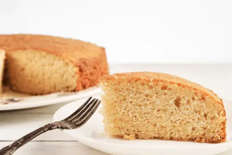 Una fetta di torta senza lievito, soffice e dorata, attende su un piatto bianco con una forchetta antica.