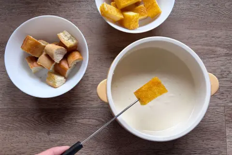 Una fetta di pane croccante viene intinta in una cremosa fonduta di parmigiano, accanto a ciotole di crostini.