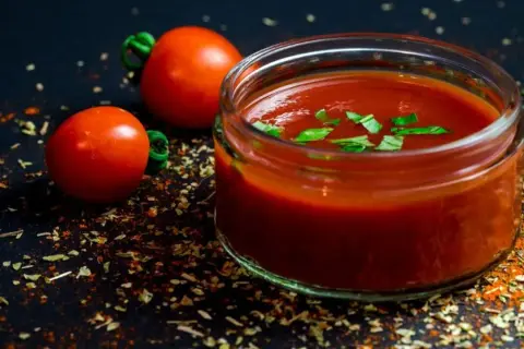 Un vasetto di sugo al pomodoro, guarnito con basilico fresco, accanto a pomodorini e spezie su sfondo scuro.
