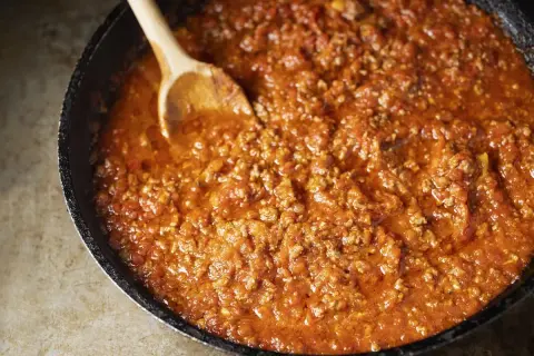 Un saporito ragù di carne in una padella, pronto per essere servito. Un cucchiaio di legno mescola la salsa rossa e densa.