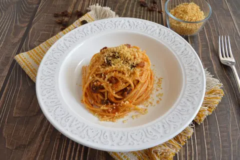 Un piatto di spaghetti con acciughe, pomodoro e pangrattato tostato, servito su un piatto bianco decorato, con un tovagliolo giallo e un bicchiere di pangrattato a lato.