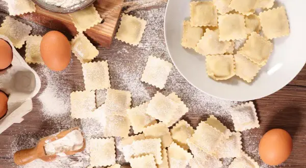 Ravioli fatti in casa sparsi su un tavolo di legno, con farina, uova e una ciotola di farina.