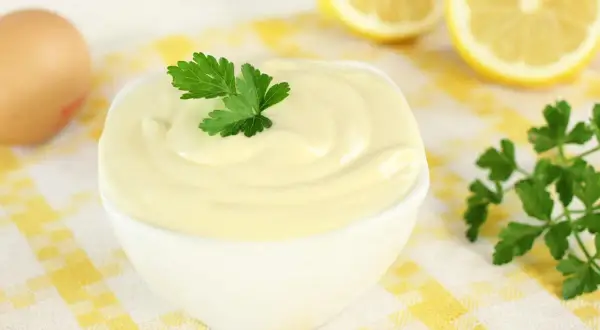 Una ciotola di cremosa maionese fatta in casa, guarnita con prezzemolo. Ingredienti freschi come uovo e limone sullo sfondo.