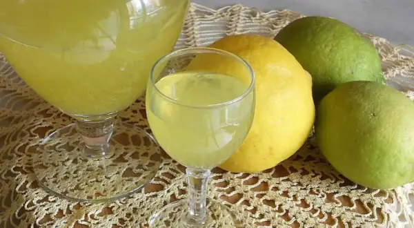 Limoncello ricetta originale Amalfi: bicchieri di liquore giallo brillante e limoni freschi, alcuni verdi, altri gialli, su un centrino di pizzo.