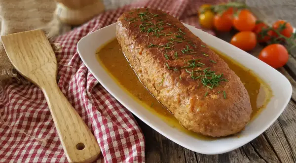 Un succulento polpettone in padella, dorato e profumato, adagiato su un letto di salsa e guarnito con erba cipollina. Accanto, una spatola in legno e pomodorini freschi.